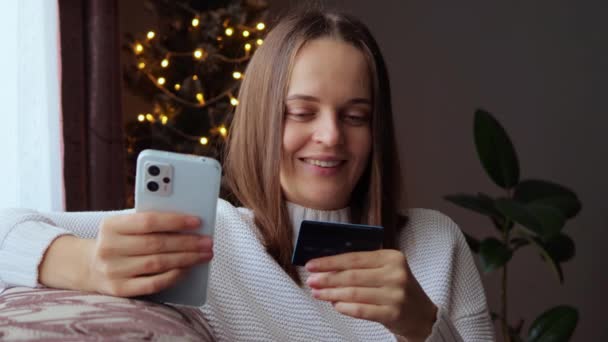 かなり見栄えの良い茶色の髪の女性は リラックスした一日を楽しんで彼女のスマートフォンを握っているクレジットカードからデータを入力し 新年の木の近くのソファに座ってオンラインショッピング — ストック動画
