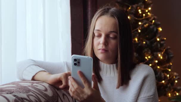 携帯電話のメッセージ オンラインセル コンフォートブラウズ デコレーションされたクリスマスツリーの近くにソファーに座って退屈なソーシャルネットワークニュースを読んでスマートフォンを使用して怒っている若い女性 — ストック動画