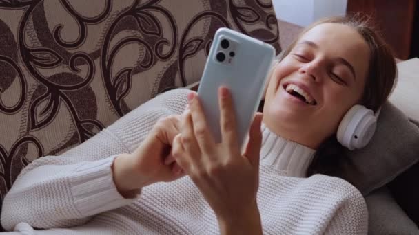ヘッドフォンのソファーに横たわる笑い女性は インターネットを閲覧したり 白いセーターを着てソーシャルネットワークで面白いビデオを見ている — ストック動画