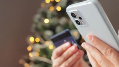 Çevrimiçi ödeme. Ev bankacılığı. Arka planda Noel ağacı ile çevrimiçi poz vermek için cep telefonu ve kredi kartı kullanan tanınmayan bir kadın.