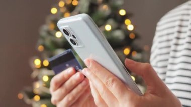 Noel zamanı kadın akıllı telefonuyla internetten tatil süslemeleri alıyor. Kredi kartı tutuyor. Kışın evde rahat otururken ödeme yapmak için veri girişi yapıyor..