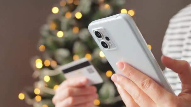 彼女のモバイルデバイスを使用して認識できない女性は 背景にクリスマスツリーで彼女の家の快適さから彼女のクレジットカードで安全なオンライン決済を行います — ストック動画