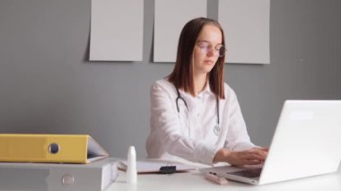 Steteskoplu, beyaz laboratuvar önlüğü giyen yorgun bir kadın. Masasında dizüstü bilgisayarla oturuyor. Bilgileri temel olarak giriyor..