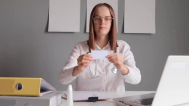 Steteskoplu kendine güvenen doktor beyaz laboratuvar önlüğü giymiş laptopuyla ofisinde oturmuş reklam için boş bir kartvizit gösteriyordu..