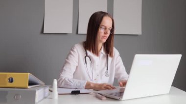 Steteskoplu, beyaz önlüklü, üzgün bir kadın doktor laptopta çalışıyor, tıbbi araştırmalar yapıyor ve bilgisayar programında sorunlar yaşıyor..