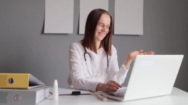 Steteskoplu, gülümseyen, beyaz önlüklü, laptopuyla ofisinde oturan ve hastasıyla hastalık tedavisi hakkında konuşurken görüntülü konuşma yapan güzel kadın doktor..