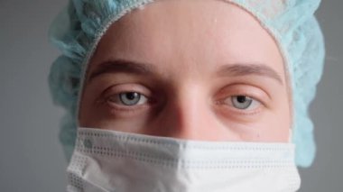 Tıbbi cerrah. Klinik tıbbı. Üniformalı tıbbi başlık takan yorgun genç bayan doktor ya da hemşire kameraya bakarak cerrahi işlemleri bitirdikten sonra maskesini çıkarıyor..