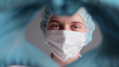 Sevimli, güler yüzlü kadın doktor ya da hemşire üniformalı, tıbbi eldiven ve maske takmış kalp şeklindeki kameraya bakıyor, aşk jestleri yapıyor, sağlık hizmetleri yapıyor..
