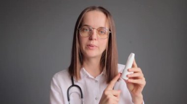 Beyaz laboratuvar önlüğü ve steteskop giymiş profesyonel bir kadın doktor. Gri arka planda izole bir şekilde duruyor. Boğaz ağrısı tedavisi için ellerinde sprey tutuyor..