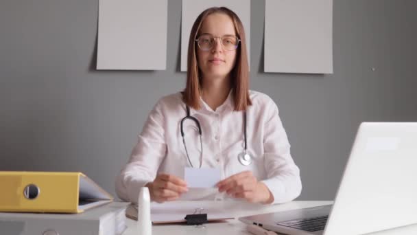 広告のための区域が付いている空の訪問カードを示すラップトップが付いているテーブルで彼女のオフィスに座っている白い実験室のコートを身に着けている笑う陽気な女性の医者 — ストック動画