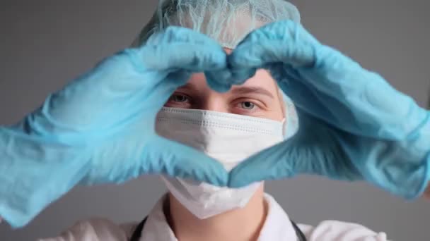 微笑着满意的女医生 心脏病医生或护士 戴着统一的医疗帽手套和面罩 双手在病房里摆出爱情的姿势 让人心花怒放 — 图库视频影像