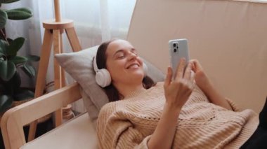 Enfes ve şık bir kadın örgü süveter giyip neşeli bir şekilde kulaklıkla müzik dinliyor evdeki kanepede şarkısını seçiyor ve müzik listesi içinde dans ediyor..