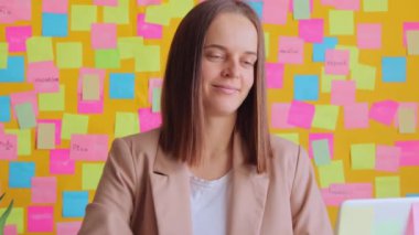 Bilgisayarda sarı duvara karşı çalışan gülümseyen bir kadın renkli çıkartmalarla işini bitirirken mutlu bir şekilde işyerinde oturuyor ve tatmin olmuş bir ifadeyle gülümsüyor..