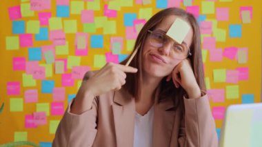 Bilgisayarda çalışan, sarı duvarlı, renkli çıkartmalı, iş yerinde sıkıcı, alınında not kartıyla duran ve gün sonuna kadar kalem tutan mutsuz bir kadın..