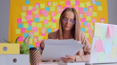 İşyerindeki dizüstü bilgisayarda çalışan ceketli ve gözlüklü konsantre bir kadın. Ofisinde çalışan çok renkli not kartlarıyla çalışıyor..