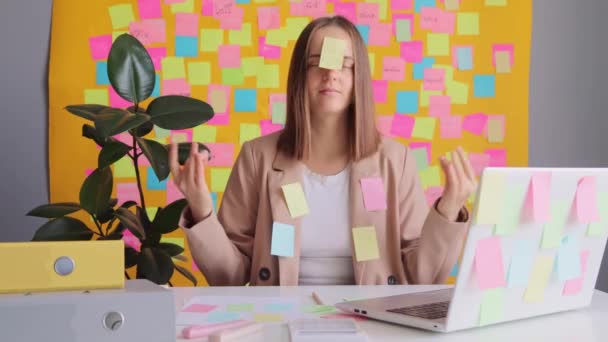疲惫的漂亮女商人穿着米黄色的夹克坐在办公室里 用笔记本电脑靠着黄色的墙壁 贴着五颜六色的贴纸 试图在工作时平静下来 沉思冥想练习瑜伽 — 图库视频影像