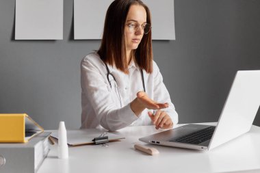 İş yerindeki ofis masasında çalışan ciddi bir doktor bilgisayarındaki monitöre bakıyor ve internet üzerinden hastasıyla konuşuyor..