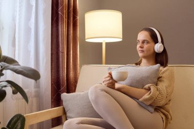 Kafkasyalı genç bir kadın, kulaklık takıp kanepede kahve ya da çayla müzik dinliyor. Sıcak içecek ve ilginç bir sesli kitap ya da podcastla eğleniyor..