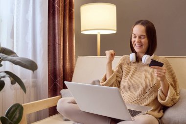 İnternetten moda alışverişi. Evde internet teknolojisi var. Sıcak süveter giyen genç bir kadın evde oturmuş başarılı alışverişi kutlarken dizüstü bilgisayar ve kredi kartıyla çalışıyor..