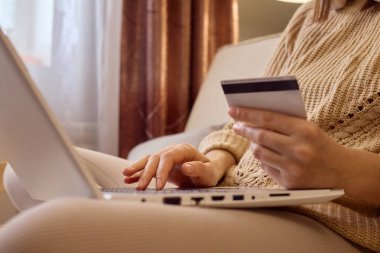 İnternetten moda alışverişi. Evde internet teknolojisi var. Sıcak süveter giyen, dizüstü bilgisayar ve kredi kartıyla çalışan, evinde koltukta oturan ve satın almak için ödeme yapan tanınmamış bir kadın..