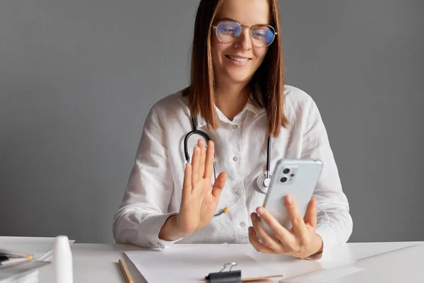Digital health specialist. Smiling woman doctor wearing white medical lab coat stethoscope and glasses using smart phone for online consultation against gray wall waving hand to her patient.