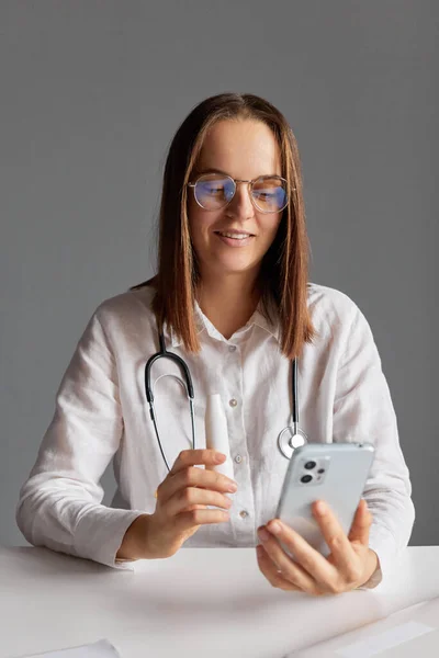 Mobile phone prescription. Woman doctor wearing white medical lab coat stethoscope and glasses using mobile phone for online consultation explaining how to use spray fore sore throat treatment.