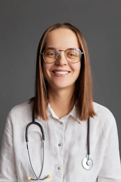 Cheerful happy professional woman doctor in glasses and white medical lab coat looking at camera sitting at workplace in clinic against gray wall smiling toothily during communicating with patient.