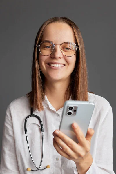 Online doctor\'s office. Digital health specialist. Mobile phone prescription. Video call with a physician. Cheerful woman doctor wearing white medical lab coat stethoscope and glasses using cell phone