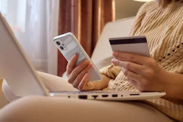 Unrecognizable woman shopping online from the comfort of home sitting on sofa with credit card mobile phone and laptop placing orders through cell phone paying in web store.