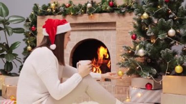 Sıcak süveter ve Noel Baba şapkası giyen mutlu bir kadın şöminenin yanında oturuyor ve güzel Noel ağacına gülümsüyor..