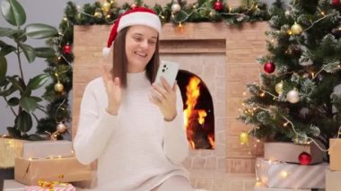 Sıcak süveter ve Noel Baba şapkası giyen arkadaş canlısı neşeli bir kadın oturma odasında şöminenin yanında oturuyor ve akıllı telefondan konuşurken Noel ağacı el sallıyor ve video görüşmesi yapıyor..