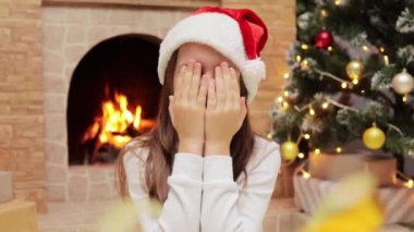 Noel Baba şapkalı küçük kız, yeni yıl hediyesini bekleyen avuçlarıyla gözlerini kapatıyor. Evde Noel ağacı ve şömineyle süslü bir odada oturuyor..