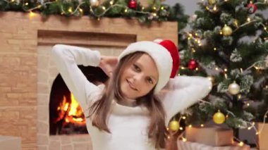 Noel Baba şapkalı neşeli küçük kız evde Noel ağacı ve şömineyle süslü bir odada oturuyor kış tatillerinin keyfini çıkarıyor ve yeni yılın tadını çıkarıyor..