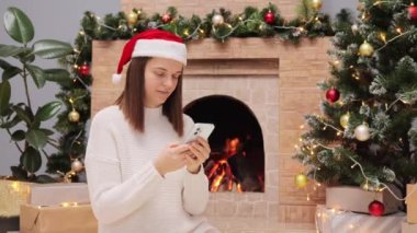 Koyu renk saçlı, kalın kazak giyen ve Noel Baba şapkalı bir kadın şöminenin yanında oturuyor ve Noel ağacı elinde cep telefonuyla şenlik fotoğraflarına bakıyor..