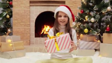 Noel Baba şapkalı neşeli küçük kız süslenmiş bir odada Noel ağacı ve şömineyle sarılıp kış tatilini kutluyor..