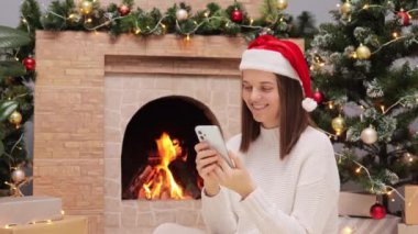 Sıcak kazak giyen neşeli ve Noel Baba şapkalı bir kadın şöminenin yanında oturuyor ve Noel ağacında akıllı telefon ekranına bakıyor..