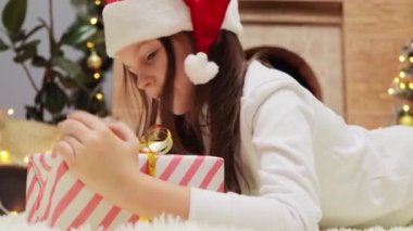 Noel Baba şapkalı, süslü bir odada Noel ağacı ve şömineyle uzanmış duran sevimli, meraklı küçük kız hediye kutusunu sallıyor..