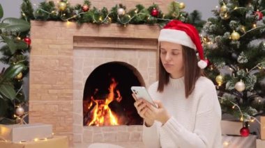 Sıcak kazak giyen üzgün kadın ve Noel Baba şapkası oturma odasında şöminenin yanında oturuyor ve Noel ağacı internetten süzülüyor canı sıkkın, neşeli bir akşam..