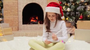 Kahverengi saçlı, kırmızı şapkalı Noel ağacı ve şöminesi olan süslü bir odada oturan Noel Baba 'ya mektup yazıp hediye listesi hazırlamasını isteyen küçük bir kız..