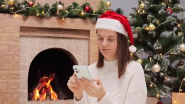 Beyaz kadın kalın kazak giyiyor ve Noel Baba şapkası takıyor şöminenin yanında oturuyor ve Noel ağacı elinde cep telefonuyla internete girip esniyor..