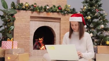 Süveter ve Noel Baba şapkası giyen yorgun bir kadın dizüstü bilgisayar kullanıyor internet işini yapıyor Noel ağacı ve şöminenin yanında halının üzerinde otururken boynu ağrıyor..