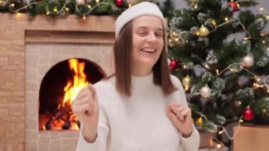 Noel Baba şapkalı neşeli kadın Noel ağacı ve şömine atmosferinin yanında dans ediyor. Oturma odasındaki dekorasyonda rahatlayan kadın..