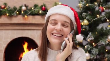 Neşeli, mutlu, cep telefonu kullanan pozitif bir kadın Noel ağacı ve şöminenin yanında oturmuş arkadaşlarıyla akıllı telefondan konuşuyor ve kış tatilini tebrik ediyor..