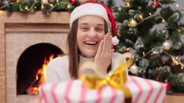 Tatil sezonunu kutluyoruz. Noel Baba şapkalı şaşkın kadın, sıcak şömine ve Noel ağacının yanında sevinç ve heyecan ifade eden hediye kutusunu memnuniyetle kabul ediyor..