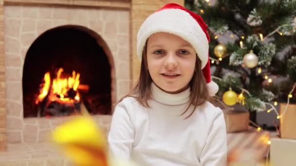 圣诞佳节前夕 身穿圣诞帽 头戴圣诞礼帽 头戴圣诞礼帽 面带微笑的小女孩与圣诞树 壁炉一起坐在装饰过的房间里 领取礼物盒 — 图库视频影像