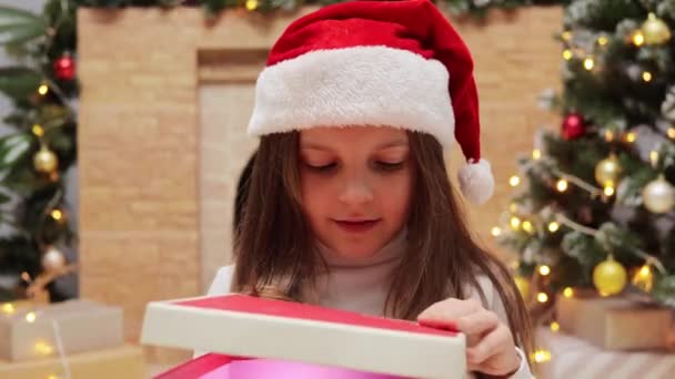 サンタクロースのハットオープンギフトボックスの中を覗くサンタクロースの小さな少女は 自宅でクリスマスツリーや暖炉で装飾された部屋に座って興奮した表情で驚きました — ストック動画