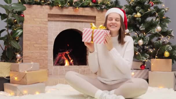 笑顔の陽気な女性はセーターを着て サンタクロースの帽子は Xmasの木と暖炉の近くに座って良いお祝いの気分の中にいることに興味を持っている現在のボックスを揺さぶります — ストック動画