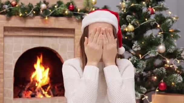 年轻女子头戴圣诞帽 身穿白色毛衣 呆在家里 围着圣诞装饰品 用手捂住眼睛 在壁炉边和圣诞树旁玩世不恭 — 图库视频影像