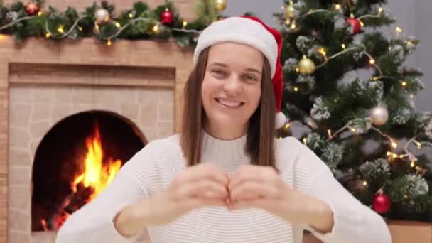 快乐的女人手牵着心形的手 戴着红红的圣诞老人帽子 在圣诞树旁的壁炉边消磨时间 看着镜头 开心地微笑着庆祝新年 — 图库视频影像