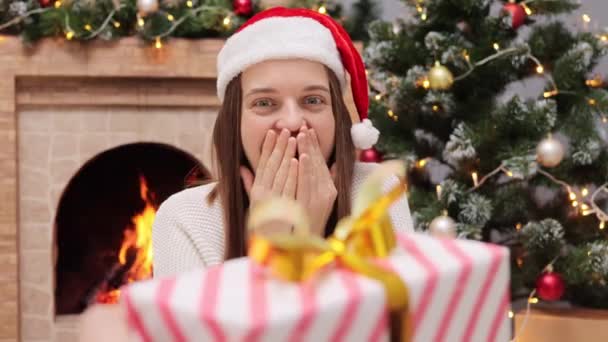 快乐的女人从男朋友那里接过礼品盒 戴着圣诞老人的帽子快乐地庆祝圣诞节和靠近壁炉的新年 看起来很惊讶 — 图库视频影像